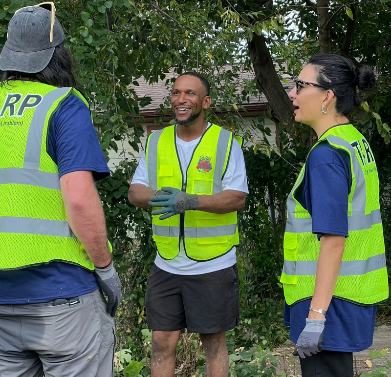 Dallas United For Good Three people in high-visibility vests standing outdoors, with a chainsaw on the ground.